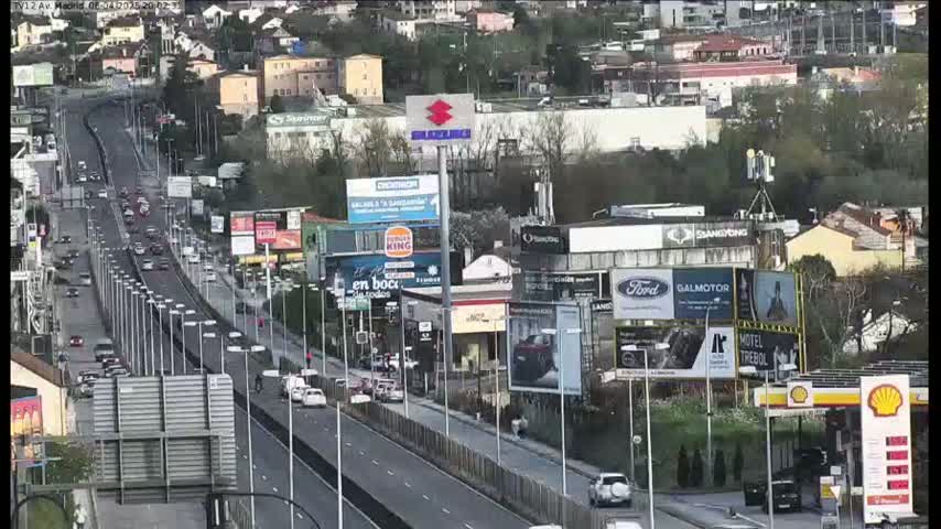 A highway with moderate traffic flows through a city with numerous billboards and a gas station, under a partly cloudy sky.