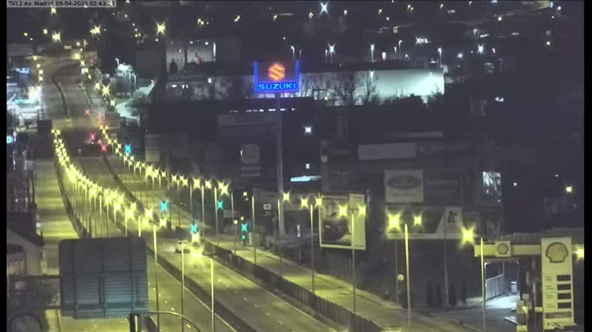 A nighttime, brightly lit highway with sparse traffic runs past a building with a Suzuki logo and other businesses.