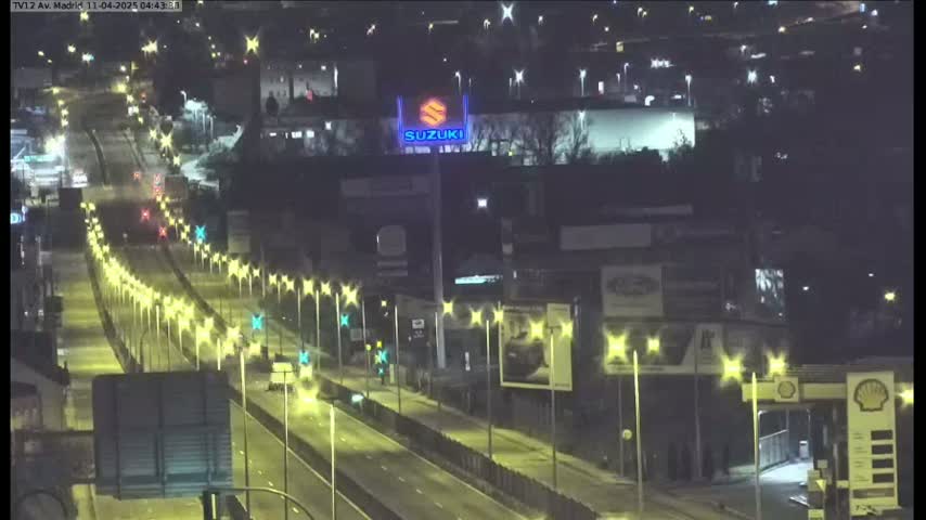 A nighttime view of a multi-lane highway with sparse traffic, lined with streetlights and bordered by buildings and a gas station.