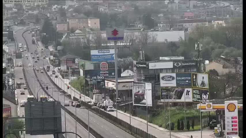 A highway scene on an overcast day shows numerous vehicles traveling along a multi-lane road, passing billboards and buildings alongside the roadway.