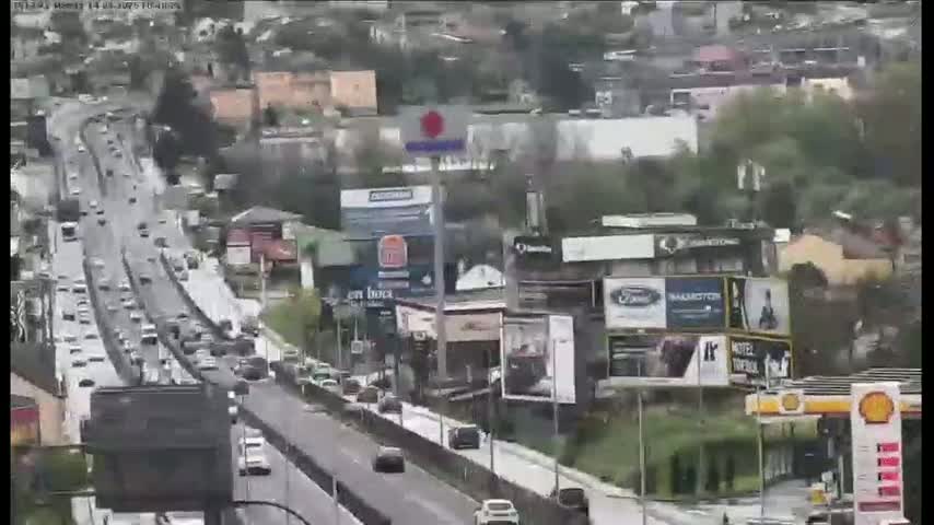 A rain-slicked highway with moderate traffic flows past buildings with numerous billboards on a hillside.