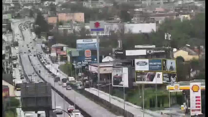 A rain-slicked highway with moderate traffic flows through a city with numerous billboards and businesses alongside it.