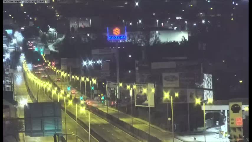 A wet, nighttime highway scene shows several lanes of traffic moving under bright streetlights past buildings with illuminated advertisements.