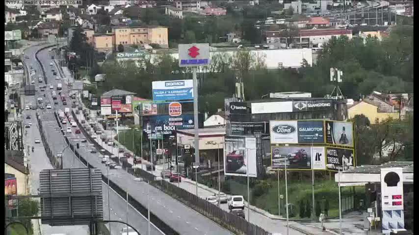 A highway scene shows numerous vehicles traveling on a multi-lane road, surrounded by buildings and billboards, under an overcast sky.