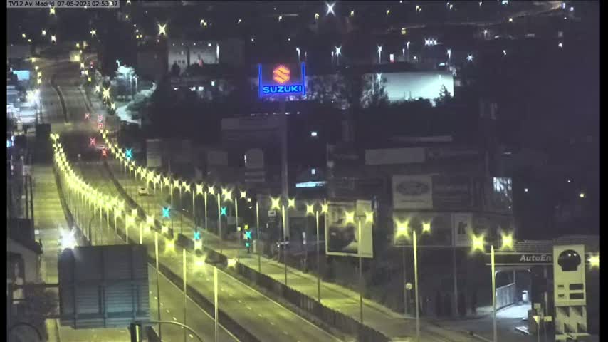 A nighttime view of a multi-lane highway with sparse traffic, lined with illuminated streetlights, alongside buildings and billboards.