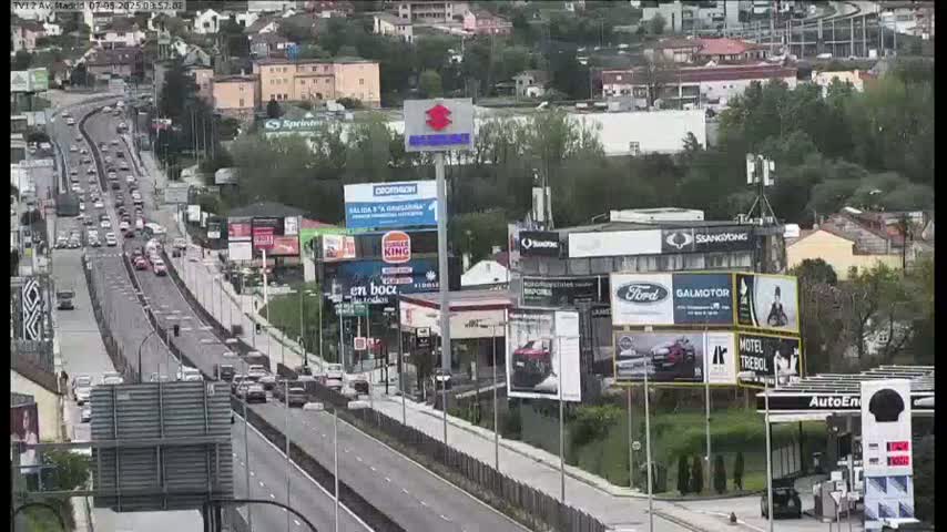 A highway with moderate traffic flows through a suburban area on a partly cloudy day, with numerous billboards and businesses lining the roadside.