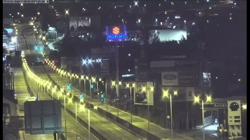 A nighttime, brightly lit highway scene shows several vehicles traveling on a multi-lane road, lined with tall lampposts, and buildings with illuminated signage in the background.