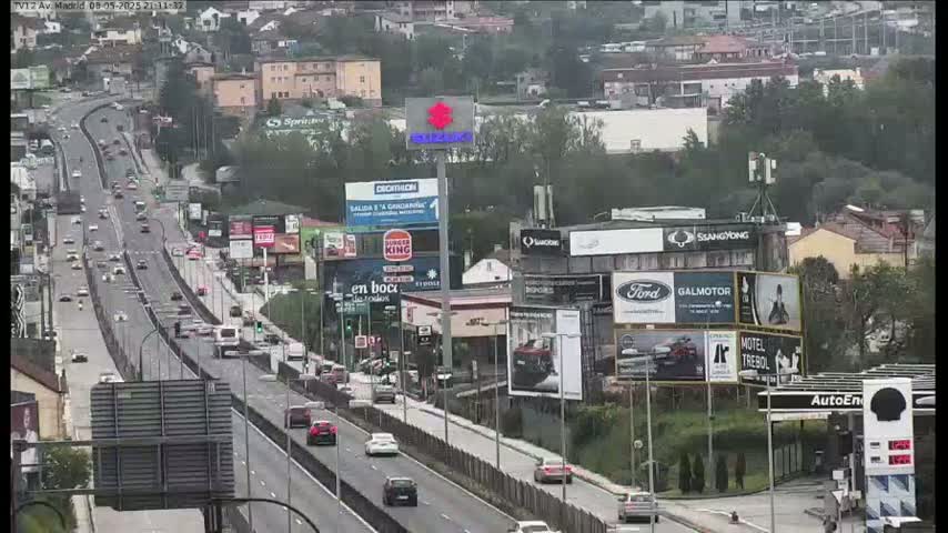 A highway scene on an overcast day shows many cars traveling in both directions, alongside billboards and buildings.