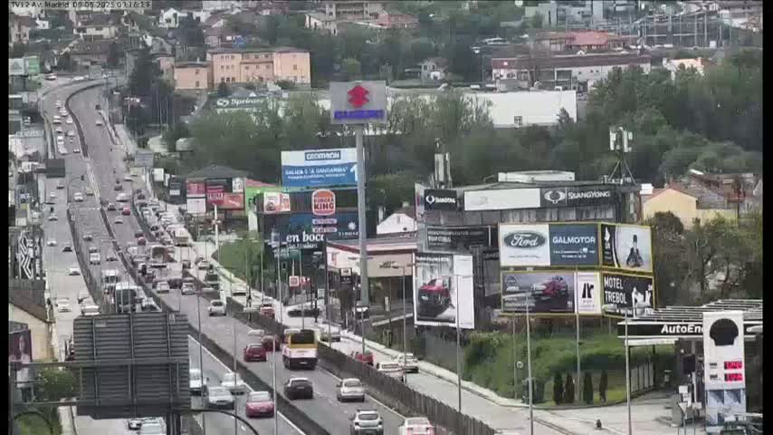 A highway with moderate traffic flows through a city on a partly cloudy day, lined with buildings and billboards.
