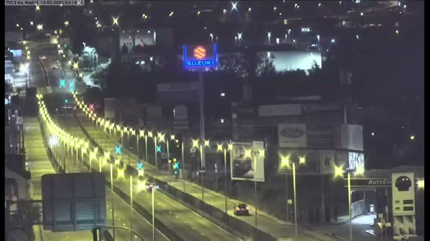 A nighttime, brightly lit highway scene shows several vehicles traveling on a multi-lane road lined with streetlights, with buildings and advertisements visible in the background.