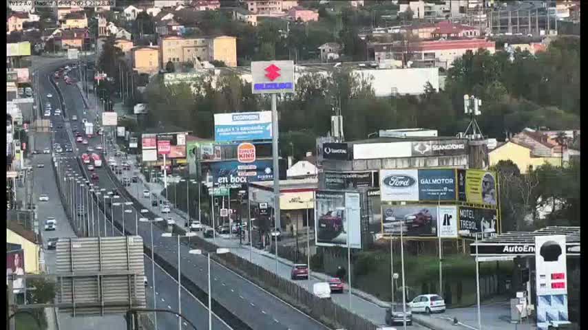 A highway scene on an overcast day shows a multi-lane highway with moderate traffic, numerous billboards and buildings lining the roadside, and cars parked and moving along adjacent roads.