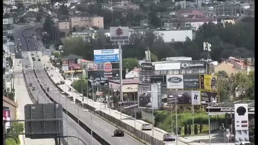A multi-lane highway with sparse traffic winds through a cityscape under a partly sunny sky, flanked by buildings with numerous billboards and a gas station.