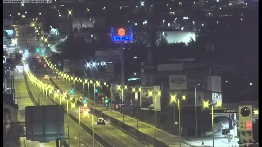 A nighttime, wet highway scene shows several vehicles traveling along a multi-lane road lined with streetlights, with buildings and a large Suzuki sign visible in the background.