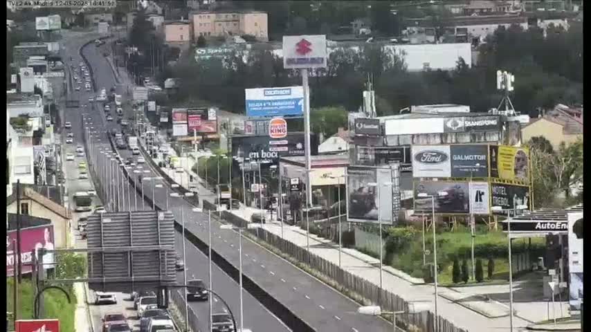A highway scene shows many cars driving on a multi-lane road beside businesses and billboards under a mostly cloudy sky.