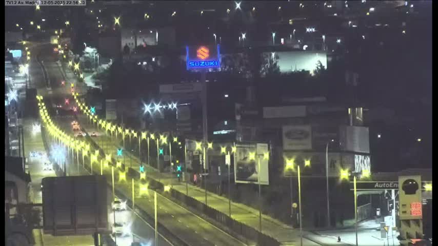A brightly lit highway at night with several vehicles driving on it, passing under streetlights and alongside buildings with billboards.