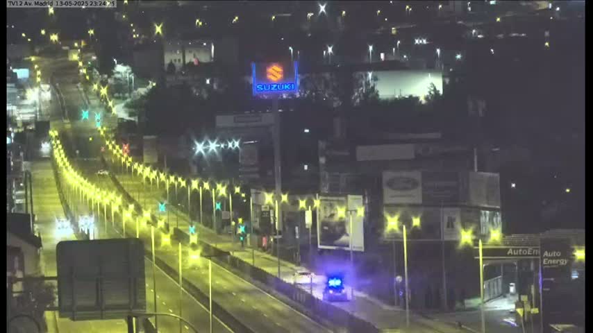 A nighttime, brightly lit highway scene shows several vehicles traveling on a multi-lane road lined with illuminated streetlights, passing by buildings with billboards.