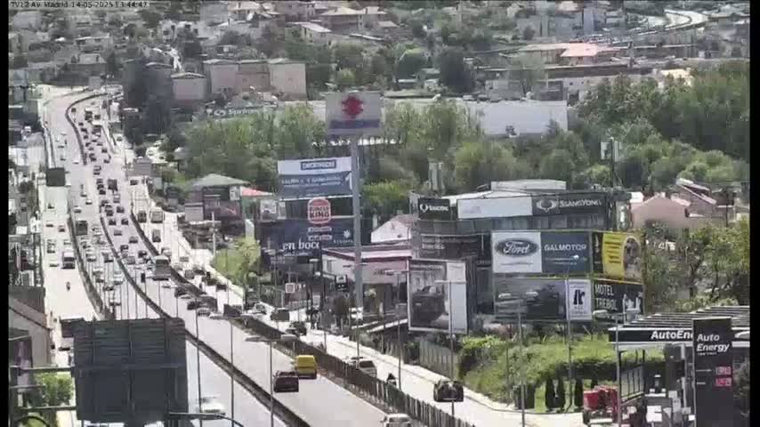 A multi-lane highway with moderate traffic flows through a suburban area under sunny skies, alongside buildings with numerous billboards.