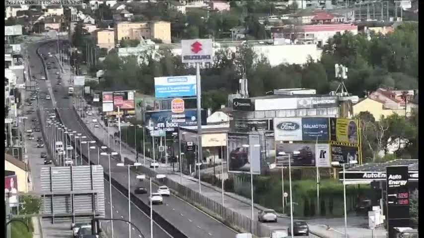 A highway with moderate traffic flows through a city on a partly cloudy day, with numerous billboards and buildings lining the roadside.