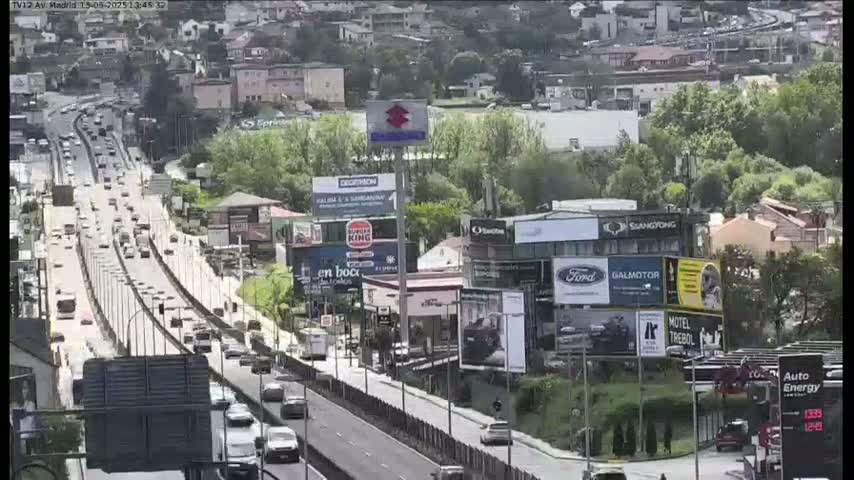 A highway with moderate traffic flows through a city on a partly cloudy day, surrounded by buildings and billboards.