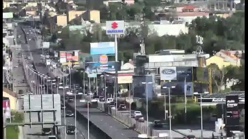 A busy highway scene shows numerous vehicles traveling on a multi-lane road surrounded by buildings and billboards, under an overcast sky.