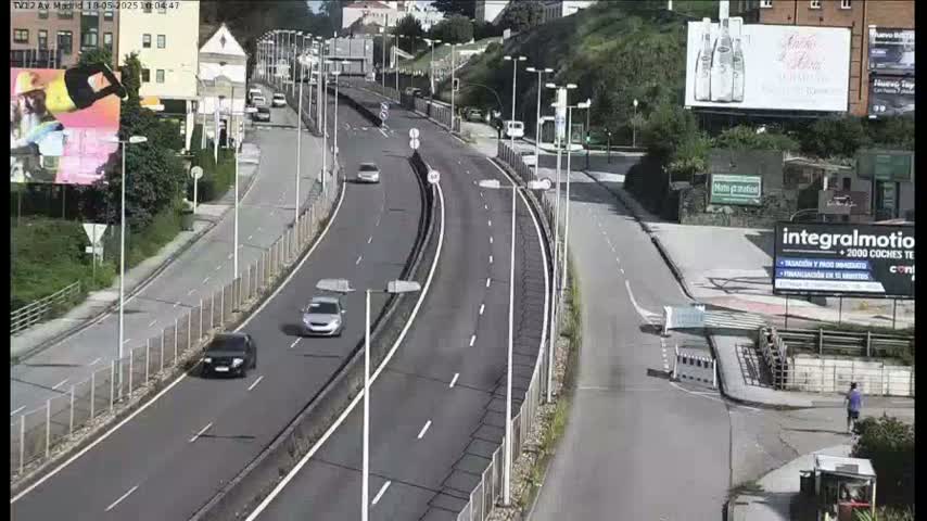 A multi-lane highway curves gently through a hilly area on a partly sunny day, with a few cars visible traveling in both directions.