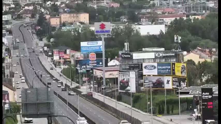 A highway with moderate traffic flows through a city under partly cloudy skies, past numerous billboards and buildings.