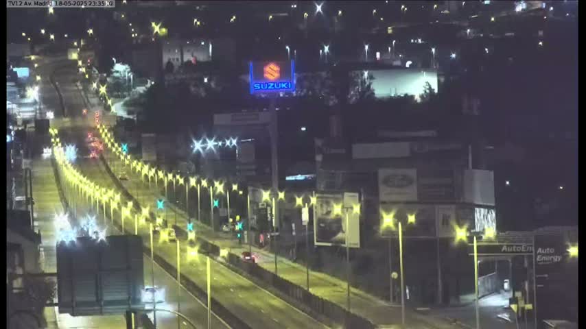 A nighttime, high-angle view of a multi-lane highway with sparse traffic, illuminated by streetlights, and passing by buildings with billboards.