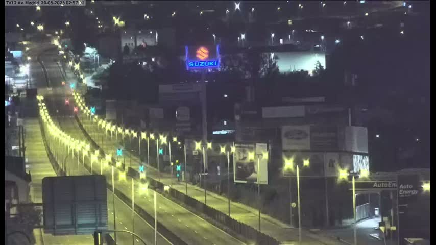 A nighttime, well-lit highway with sparse traffic passes by buildings and a large Suzuki sign.