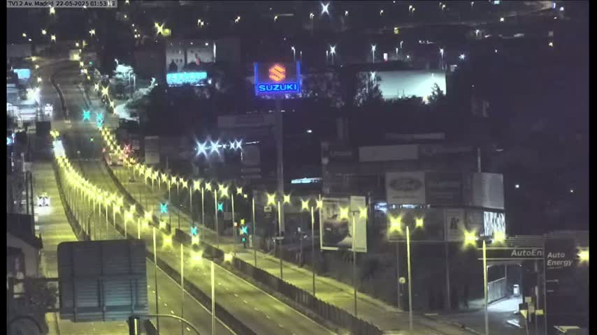 A brightly lit highway at night with sparse traffic, lined with streetlights, and buildings visible in the background under a clear sky.