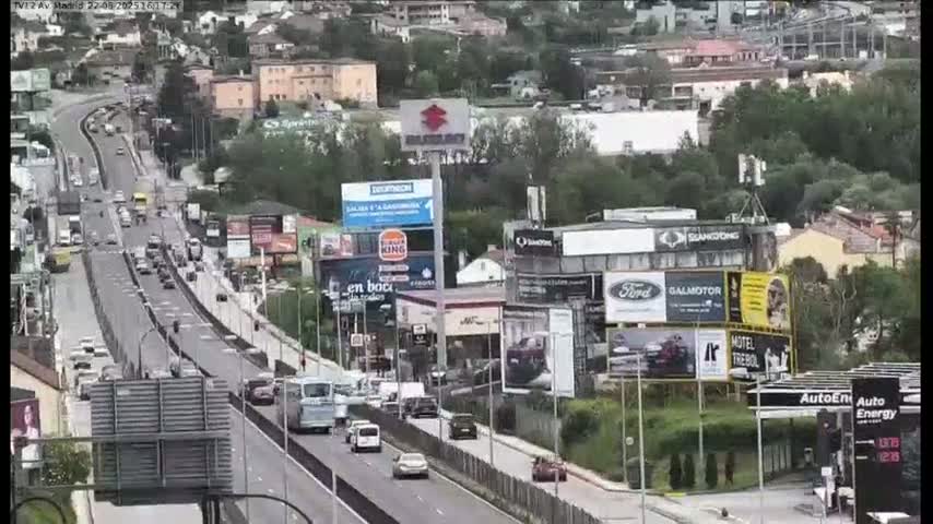 A highway scene on a partly cloudy day shows numerous vehicles traveling along a multi-lane road, passing by buildings with various billboards and advertisements.