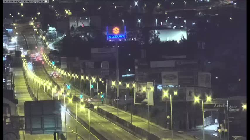 A nighttime view of a multi-lane highway with sparse traffic, lined with illuminated lampposts, and buildings with billboards visible in the background.