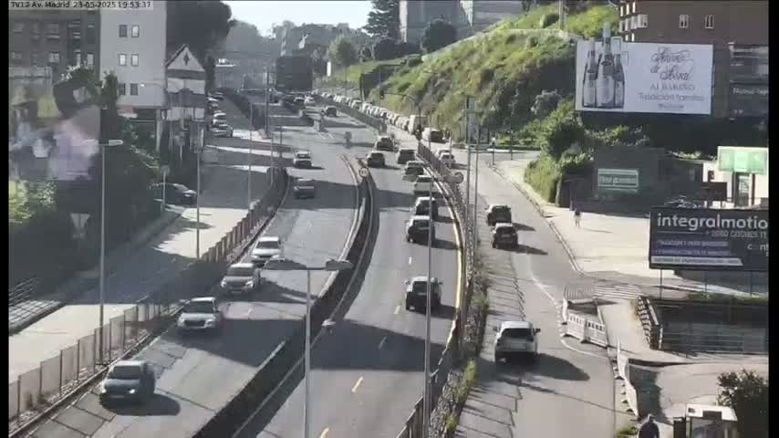 A sunny day on a multi-lane highway with moderate traffic flowing alongside buildings and green hills.
