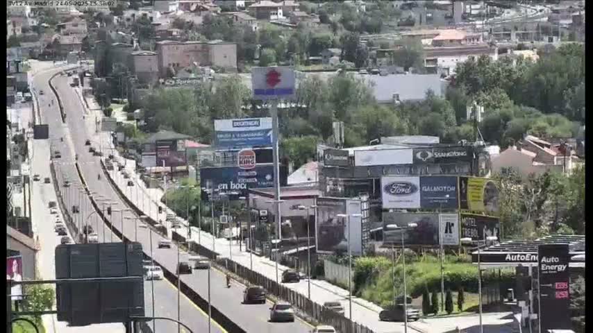A multi-lane highway with moderate traffic flows through a hilly area with buildings and billboards on a sunny day.