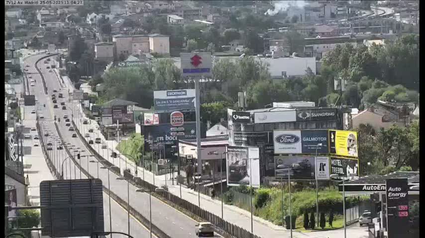 A highway with moderate traffic flows through a city on a sunny day, bordered by buildings, billboards, and lush green vegetation.
