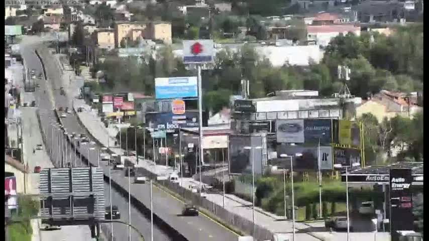 A multi-lane highway with moderate traffic flows through a city under a partly sunny sky, past numerous businesses and billboards.