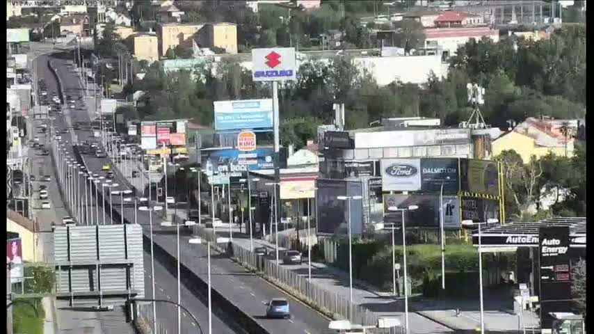 A highway scene on a sunny day shows numerous vehicles driving along a multi-lane road lined with billboards and businesses.