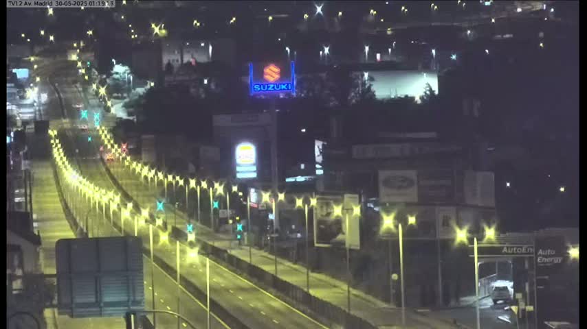 A nighttime, wet highway scene shows several vehicles driving under illuminated streetlights alongside commercial buildings.