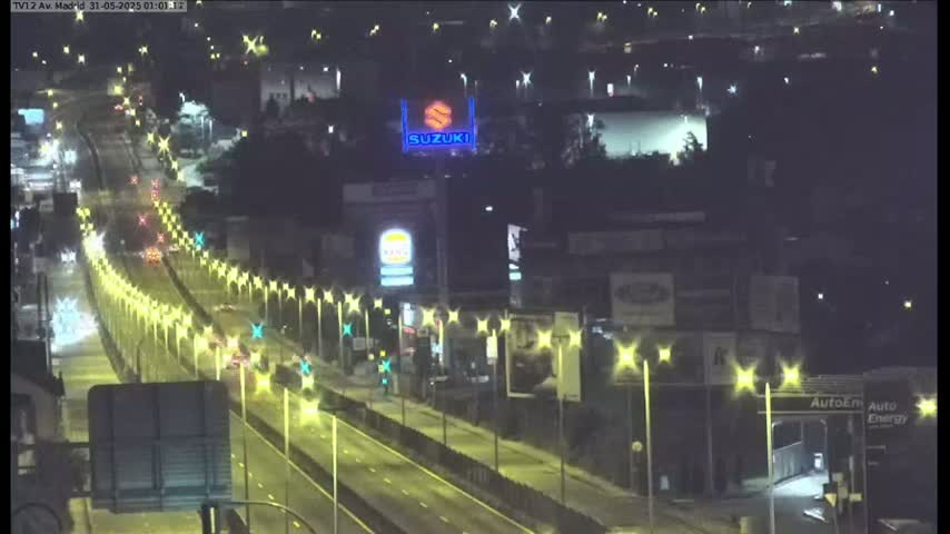 A nighttime, high-angle view shows a multi-lane highway with sparse traffic illuminated by streetlights, bordered by buildings and billboards.
