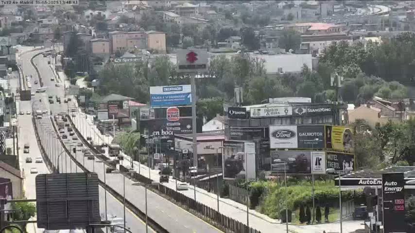 A highway with moderate traffic flows through a city under partly cloudy skies, passing by numerous billboards and buildings.