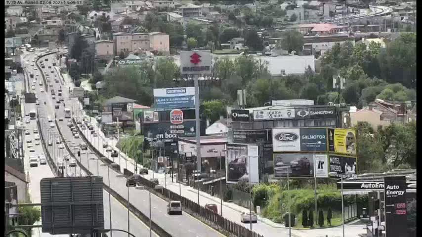 A highway with moderate traffic flows through a city on a partly cloudy day, surrounded by buildings and billboards.
