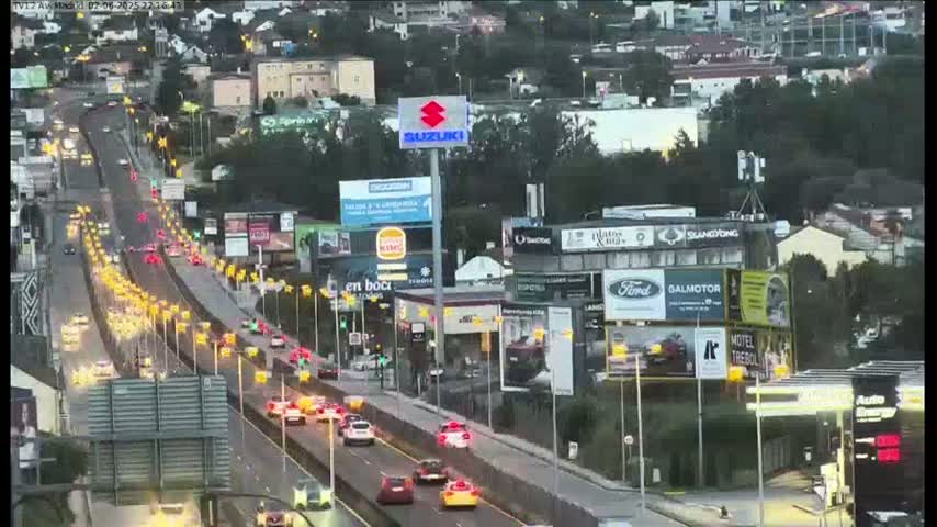 A busy highway with multiple lanes of traffic flowing in both directions, under overcast skies, passes by billboards and buildings.