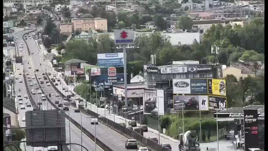 A highway with moderate traffic flows past businesses and billboards on a partly cloudy day.