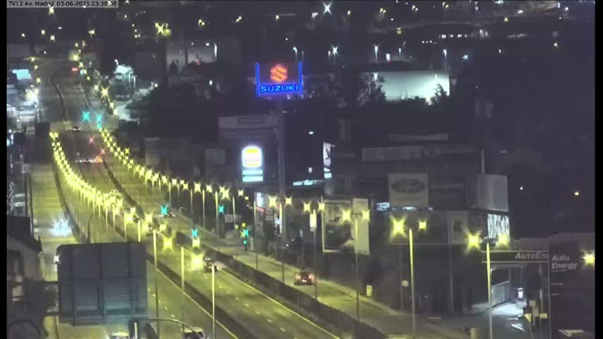A nighttime, brightly-lit highway scene shows several vehicles traveling along a multi-lane road, lined with streetlights and surrounded by buildings and businesses.