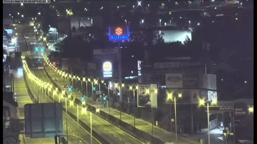 A nighttime, well-lit highway scene shows several cars driving on a multi-lane road, lined with streetlights, passing by various businesses and billboards.
