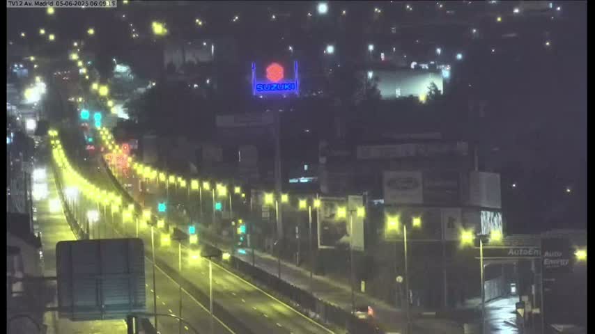 A rain-slicked highway at night is illuminated by streetlights, showing light traffic and a large illuminated Suzuki sign in the background.