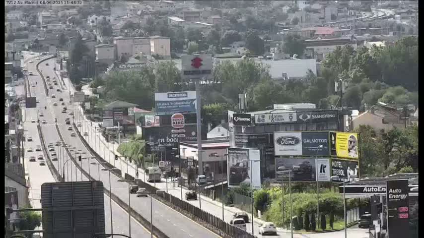 A highway with moderate traffic flows through a city on a partly cloudy day, passing numerous billboards and buildings.