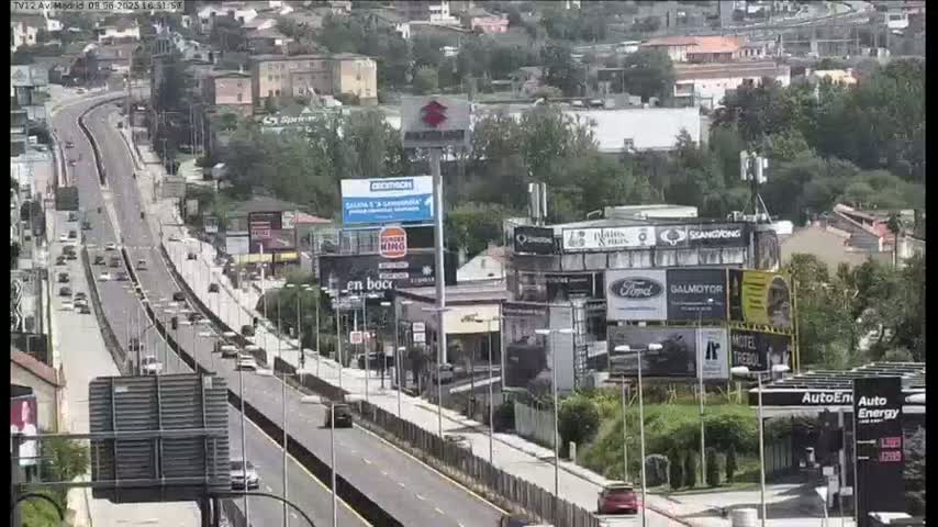 A highway with moderate traffic flows through a city under a partly cloudy sky, passing numerous buildings and billboards.