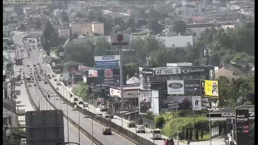 A highway with moderate traffic flows through a city on a partly sunny day, with numerous billboards visible along the roadside and buildings flanking the highway.