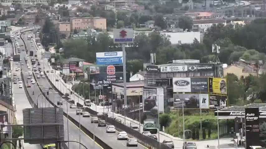 A multi-lane highway with moderate traffic flows through a city on a partly cloudy day, surrounded by buildings and billboards.