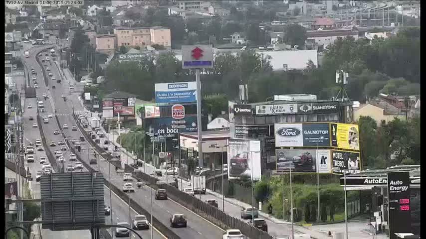 A highway with moderate traffic flows past buildings with numerous billboards and advertisements, under a partly cloudy sky.